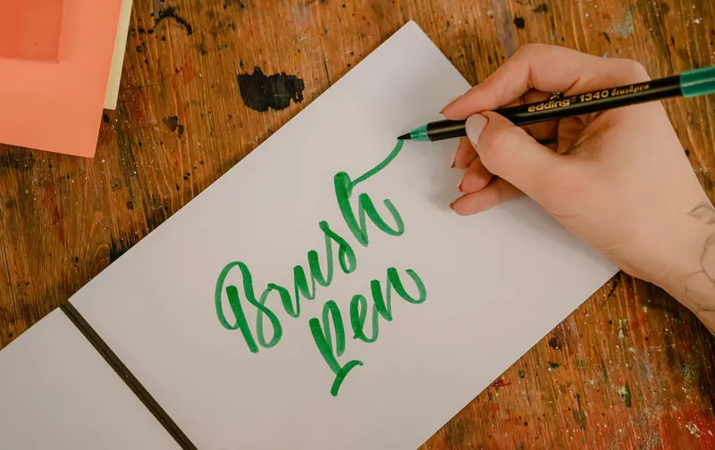 Top view of crop anonymous female designer writing BRUSH PEN title using felt pen and creative letter font while sitting at wooden table with ink spots on surfa
