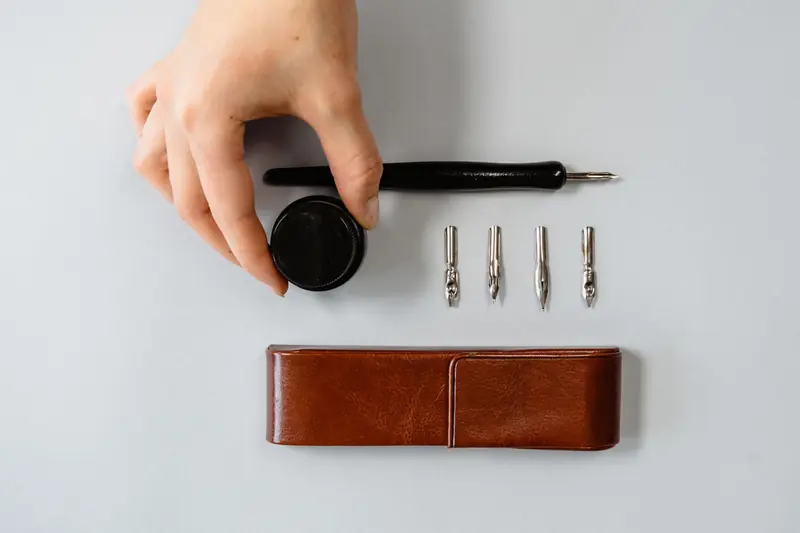 A hand arranging calligraphy tools, including nibs, ink, and pen on a white background