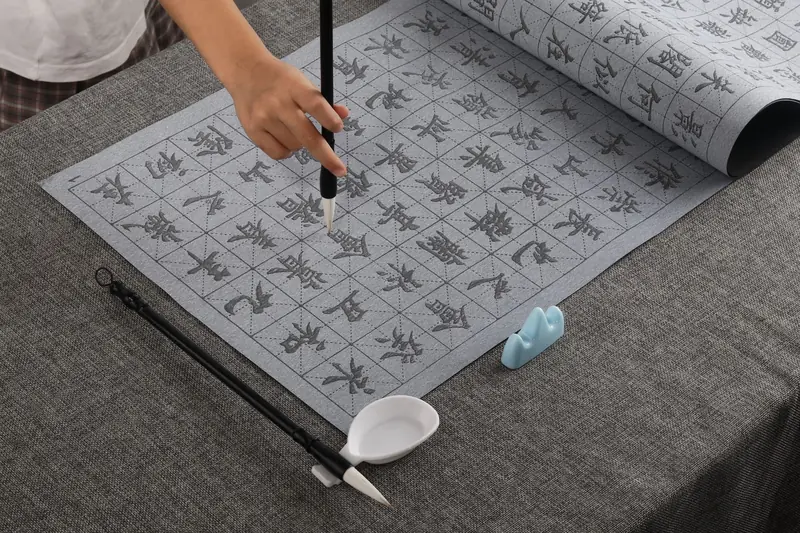 A hand demonstrates calligraphy techniques using a brush on grid paper, showcasing traditional art