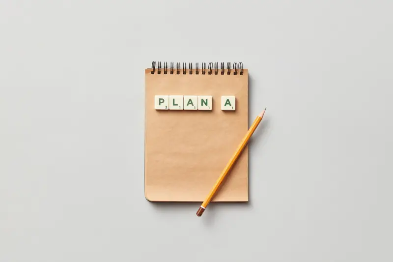 Top view of a notepad with a pencil and scrabble tiles spelling 'PLAN A' on a gray background