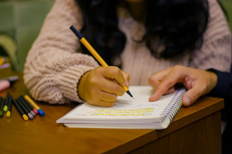 A person writing notes in a notebook with colorful pens on a wooden desk